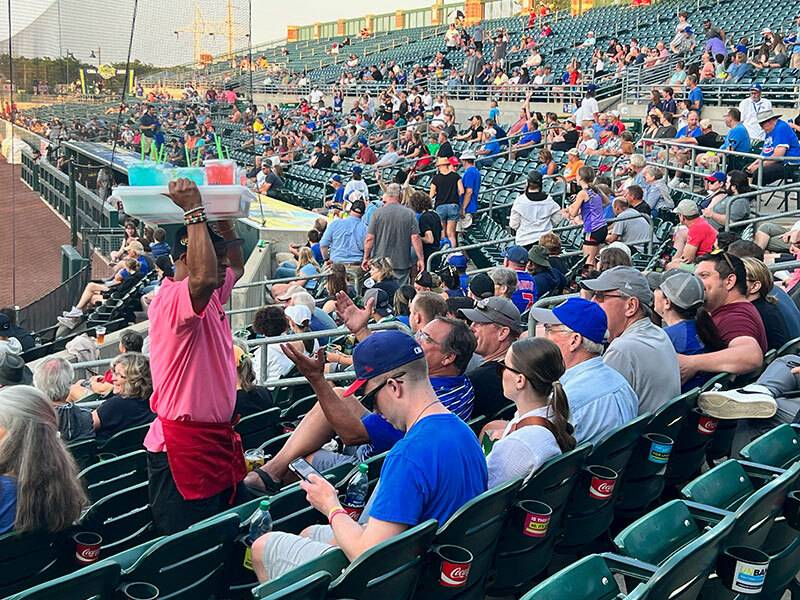 Man in pink t-shirt and red apron with tray of flavored lemonades over his head standing in stadium seats with people watching a baseball game