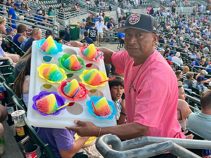 Man in pink t-shirt and ball cap standing in stadium seats at a ball holding a tray full of sno-cones